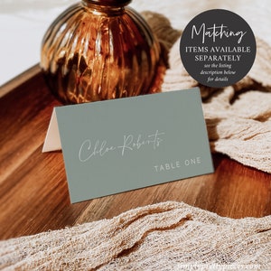May include: A sage green place card with the name "Chloe Roberts" written in white script and the text "TABLE ONE" printed in white below the name.