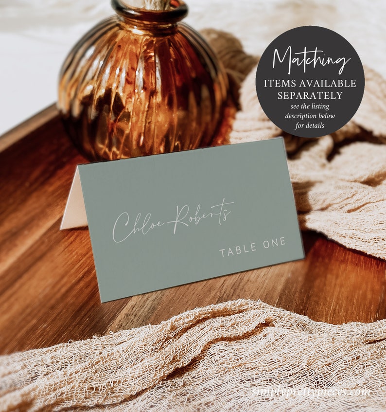 May include: A sage green place card with the name "Chloe Roberts" written in white script and the text "TABLE ONE" printed in white below the name.