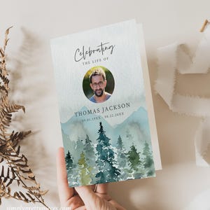 May include: A watercolor memorial card with a photo of a man in a circle. The card has a blue and green watercolor background with mountains and trees. The text reads "Celebrating the Life of Thomas Jackson 09.01.19XX - 08.22.20XX".