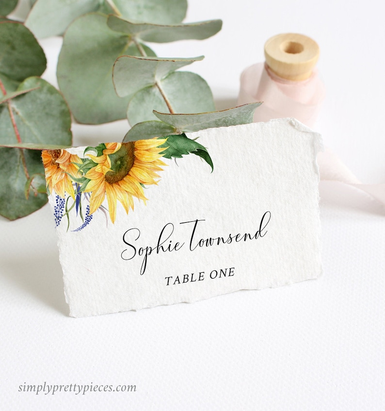 May include: A white place card with a watercolor sunflower design. The text "Sophie Townsend" and "Table One" is printed in black.