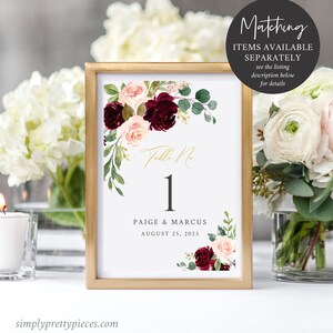 May include: A framed wedding table number card with a gold border. The card features the number 1 in black, the names Paige & Marcus, and the date August 25, 2023. Floral accents include burgundy and blush roses with green foliage. The text "Matching items available separately" is also visible.