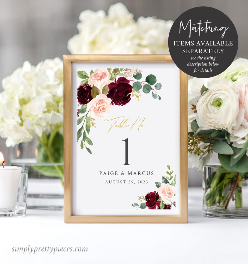 May include: A framed wedding table number card with a gold border. The card features the number 1 in black, the names Paige & Marcus, and the date August 25, 2023. Floral accents include burgundy and blush roses with green foliage. The text "Matching items available separately" is also visible.