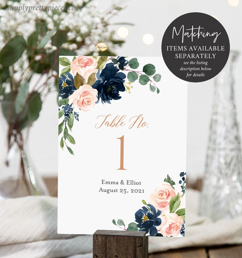 May include: A white table number card with a floral design in shades of pink and navy blue. The card has the text "Table No. 1" in gold lettering and the text "Emma & Elliot August 25, 2021" in black lettering.