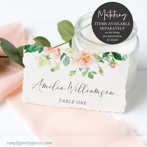 Op de afbeelding: Een wit placecard met een aquarel bloemenontwerp in roze en groene tinten. De tekst "Amelia Williamson" en "Tafel E&eacute;n" is in grijs op de kaart gedrukt.