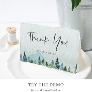 Puede incluir: Una tarjeta de agradecimiento con una ilustraci&oacute;n de acuarela de una cordillera y pinos. El texto "Gracias" est&aacute; escrito en letra cursiva negra y los nombres "Helen & Elliot" est&aacute;n impresos debajo.
