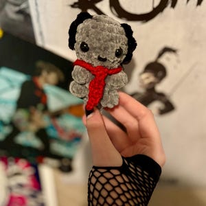 May include: A handmade gray crochet dog with black ears and a red scarf. The plush toy has black button eyes and is held in front of a blurred background. The dog is approximately 4 inches tall.