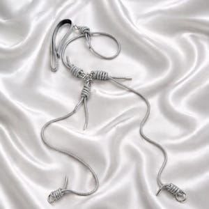 May include: A silver-colored necklace with a unique design. The necklace features a series of knots and a triangular pendant. The necklace is displayed on a white satin background. The necklace has a hook clasp and a loop.