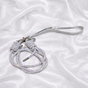 May include: A silver-colored leash with a loop handle and a decorative knot design. The leash is covered in sparkling rhinestones, and the background is a white satin fabric.