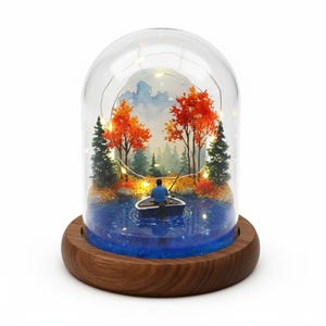 May include: A miniature diorama scene within a glass dome. It shows a person fishing from a boat on a blue lake, with trees displaying red and green leaves. The dome rests on a wooden base, and fairy lights are strung inside the dome.