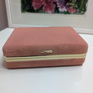 May include: A rectangular jewelry box covered in dusty pink velvet fabric. The box features a gold-toned metal trim along the center seam and a small, rectangular gold-toned metal clasp centered on the front edge for secure closure.