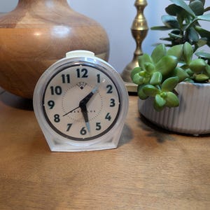 May include: A white Westclox alarm clock with a round face, black Arabic numerals, and black hour and minute hands. The clock features a light blue second hand and a small alarm indicator hand. The casing is made of white plastic with a clear cover.