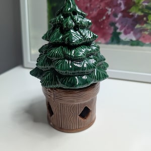 May include: A glossy ceramic figurine shaped like a dark green pine tree with textured branches, resting on a brown cylindrical base designed to resemble wood grain, featuring small diamond-shaped cutouts around the lower perimeter.