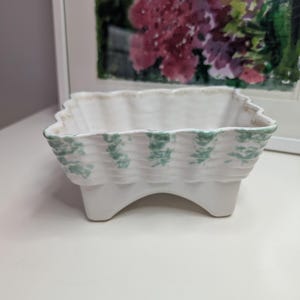 May include: A white ceramic planter with a scalloped rim and three small feet. The planter has a woven texture with a green floral pattern along the rim. The background includes a framed watercolor painting.