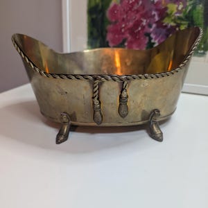 May include: An oval brass bowl with a polished finish, featuring a decorative rope-style border along the top edge and two hanging tassel accents on the front. The bowl is supported by four small, curved feet that elevate it from the surface.