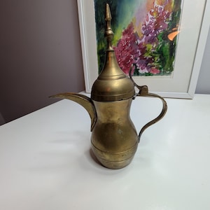 May include: A brass dallah coffee pot featuring a bulbous body, a long curved spout, a hinged lid with a pointed finial, and a curved handle. The metal surface shows a muted gold tone with subtle signs of wear and natural oxidation.