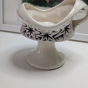 May include: White ceramic gravy boat with a pedestal base and a handle. The boat features a black starburst pattern around the middle. The interior is a cream color. The gravy boat is on a white surface.
