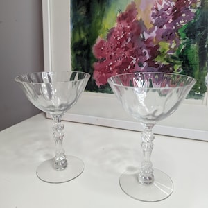 May include: Two clear, vintage champagne coupe glasses. The glasses have a wide, shallow bowl and a decorative stem with a round base. The background features a framed watercolor painting with purple and green hues.