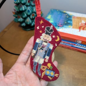 May include: A small, burgundy-colored needlepoint stocking ornament featuring a nutcracker figure in a blue and white uniform. The design includes a yellow crescent moon, stars, and a gift box at the bottom. It has a red fabric hanging loop.