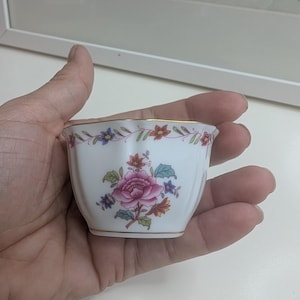 May include: A small, white teacup with a floral design. The cup features a pink rose and other colorful flowers. The rim and base are trimmed with gold. The cup is held in a person's hand.