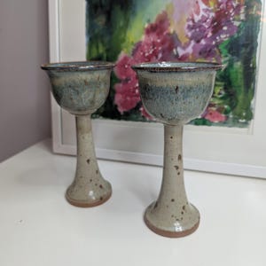 May include: Two handmade ceramic goblets. The goblets have a speckled beige stem and base, with a flared bowl in shades of blue and brown. The rim of the bowl is a darker brown. The goblets are displayed on a white surface.