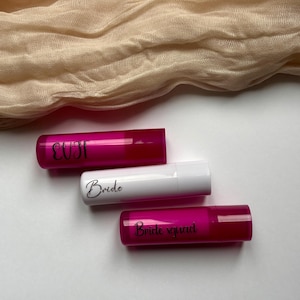Peut inclure: Trois tubes de baume à lèvres. Deux sont rose vif, un est blanc. Les tubes roses portent les mots "EUST" et "Bride squad". Le tube blanc porte le mot "Bride". Un tissu beige est en arrière-plan.