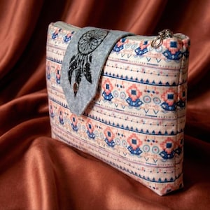 Puede incluir: Un estuche de tela rectangular con un patrón horizontal repetitivo de formas geométricas en tonos melocotón, azul y blanco. Una solapa de fieltro gris está unida al frente, mostrando un diseño de atrapasueños bordado en negro.