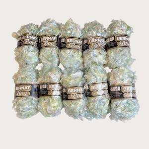 May include: Ten skeins of Bernat Boa yarn arranged in two rows of five. Each skein features a pale, iridescent green and white feathery texture with a black paper label wrapped around the center displaying the brand name Bernat and Boa.