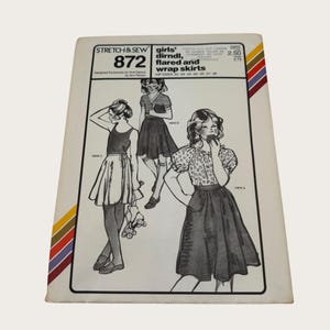 May include: Vintage sewing pattern titled "STRETCH & SEW 872" for girls' dirndl, flared, and wrap skirts. The cover features illustrations of the skirt designs. The pattern includes hip sizes 22-28. The cover has a colorful stripe design on the side.