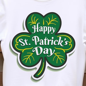 May include: White t-shirt with a green shamrock design and the text "Happy St. Patrick's Day" in white. The shamrock has yellow accents and a black outline. The shirt is displayed on a wooden hanger.