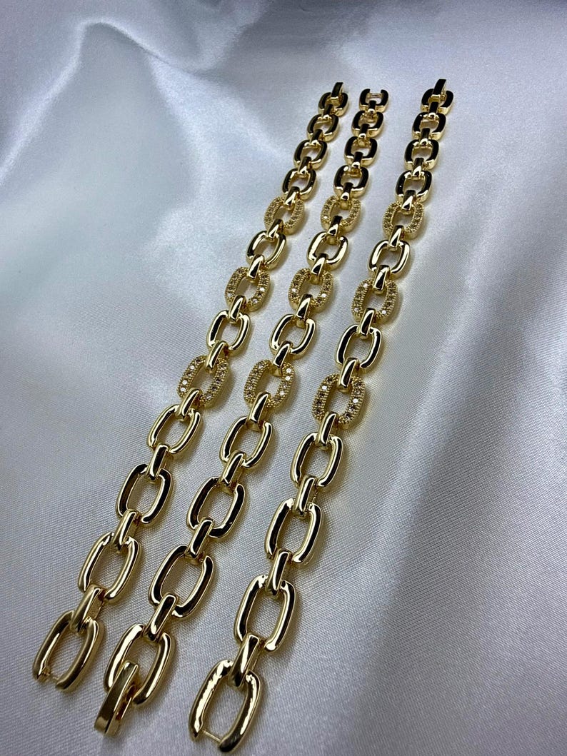 May include: Three gold-colored chain bracelets with a polished finish. Each bracelet features a series of elongated oval links, with some links adorned with small, sparkling embellishments. The bracelets are displayed on a white, satin-like surface.