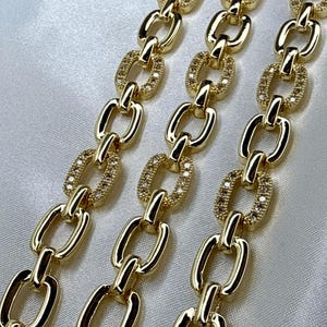 May include: Close-up of three gold-colored chain necklaces. Each chain link alternates between a solid oval and an oval encrusted with small, clear stones. The chains are displayed on a white, satin-like surface.