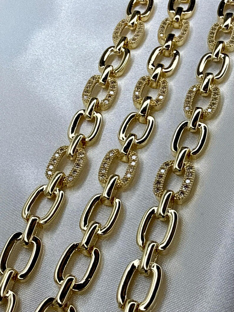 May include: Close-up of three gold-colored chain necklaces. Each chain link alternates between a solid oval and an oval encrusted with small, clear stones. The chains are displayed on a white, satin-like surface.