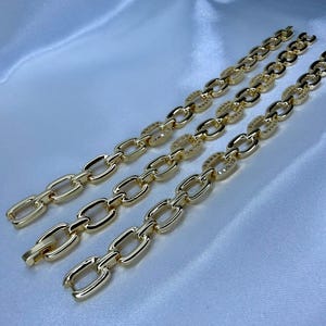 May include: Three gold-colored chain bracelets with oval links, some encrusted with small, sparkling stones. The bracelets are displayed on a white, silky fabric, creating a luxurious aesthetic. The chains are connected with a clasp.