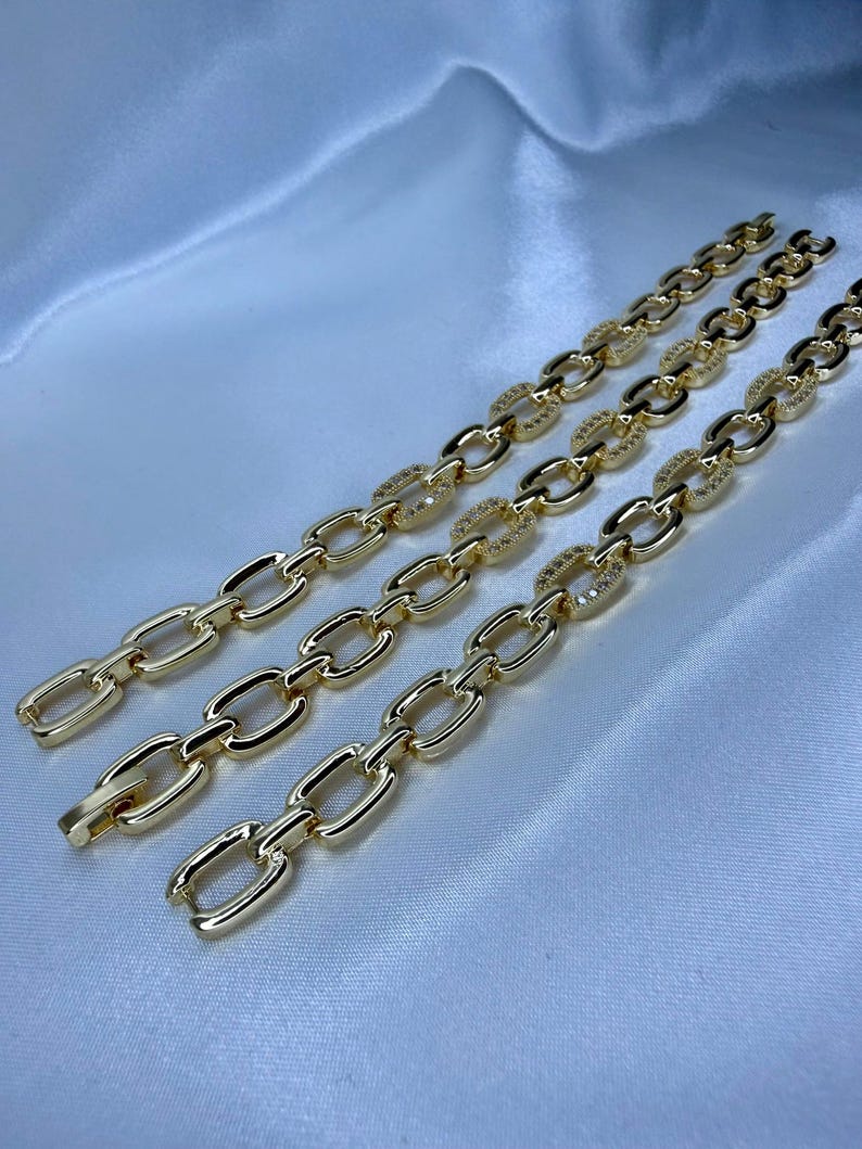 May include: Three gold-colored chain bracelets with oval links, some encrusted with small, sparkling stones. The bracelets are displayed on a white, silky fabric, creating a luxurious aesthetic. The chains are connected with a clasp.