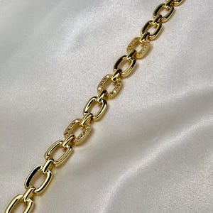 May include: A gold-colored chain bracelet with oval links. Some links are encrusted with small, sparkling stones. The bracelet is displayed on a white, silky surface, creating a luxurious aesthetic.