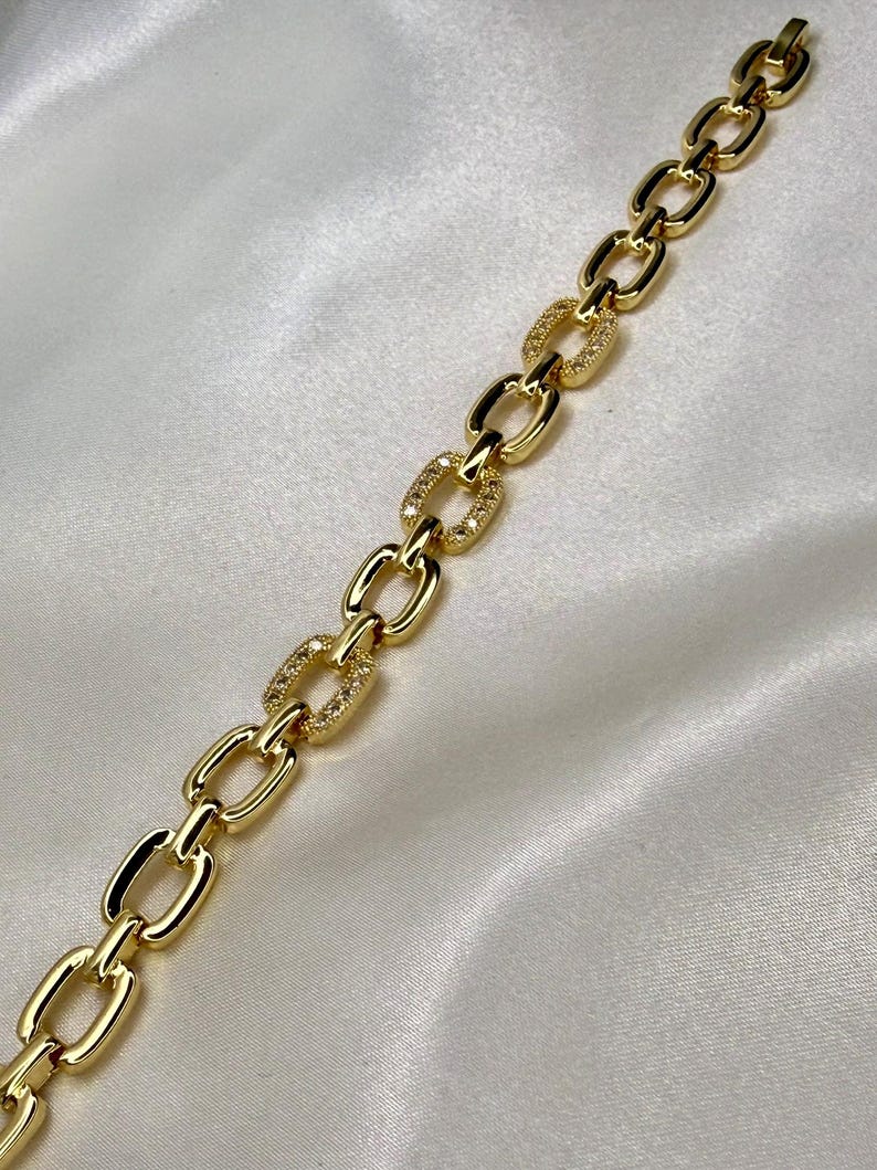 May include: A gold-colored chain bracelet with oval links. Some links are encrusted with small, sparkling stones. The bracelet is displayed on a white, silky surface, creating a luxurious aesthetic.