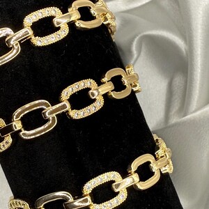 Könnte beinhalten: Goldfarbene Kettenarmbänder mit einem Kettenglied-Design. Einige Glieder sind mit kleinen, klaren Edelsteinen besetzt. Die Armbänder werden auf einer schwarzen Oberfläche mit einem weißen und grauen Hintergrund präsentiert.