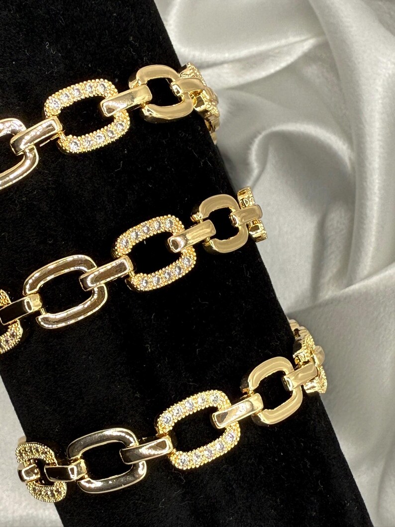 May include: Gold-toned chain bracelets with a link design. Some links are encrusted with small, clear gemstones. The bracelets are displayed on a black surface with a white and gray background.