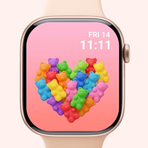 Op de afbeelding: Een gouden Apple Watch met een roze band toont een scherm met een hart van kleurrijke gummyberen. Het scherm toont ook de datum, vrijdag de 14e, en de tijd, 11:11 uur.