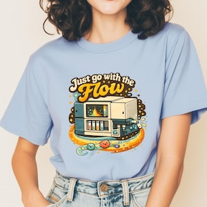 May include: Pale blue t-shirt featuring the text "Just go with the Flow" in yellow and orange. The graphic shows a retro-style machine with test tubes, a rainbow, and planets. The shirt is made of a soft material.