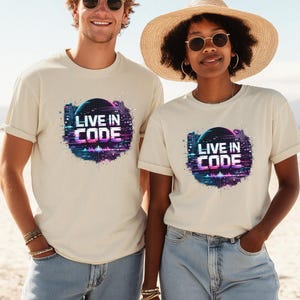 Könnte beinhalten: Zwei Personen tragen cremefarbene T-Shirts mit dem Text "LIVE IN CODE" in einem futuristischen Design. Die Grafik zeigt einen digitalen Globus und eine Stadtlandschaft in Blau-, Lila- und Rosatönen. Beide tragen Sonnenbrillen und Jeans.