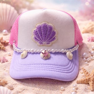 May include: A trucker hat with a white front panel, pink mesh back, and a lavender brim. The hat features a purple seashell design and is adorned with a pearl and seashell chain. The hat is decorated with a small pink starfish.