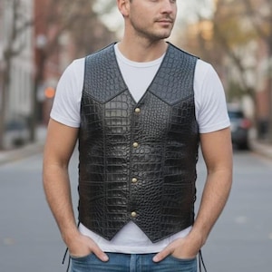 Peut inclure: Gilet en cuir noir avec une texture peau de crocodile, col en V et boutons dorés. Le gilet est porté sur un t-shirt blanc et associé à un jean bleu. Le gilet a des lacets latéraux.
