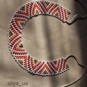 Puede incluir: Collar de cuentas hecho a mano con un patrón geométrico. El collar presenta cuentas naranjas, blancas, rojas y doradas. El diseño incluye patrones de galones y triángulos. El collar está sobre un fondo beige y tiene lazos de cuerda blanca.