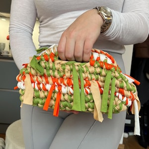 May include: A handmade clutch bag with a woven design. The bag features a mix of orange, green, white, and beige fabric strips. The clutch has a rectangular shape with a handle and decorative fringe.