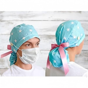 Puede incluir: Un gorro quirúrgico azul claro con un estampado de estrellas blancas y lazos de cinta rosa. El gorro tiene un botón en el lateral para las correas de la mascarilla. La persona lleva una camiseta blanca y una mascarilla quirúrgica.