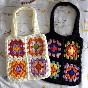 Puede incluir: Dos bolsos de mano de ganchillo. Uno es de color crema, el otro negro. Cada bolso presenta un patrón cuadrado con diseños florales coloridos en tonos naranja, morado, rosa, azul y verde. Los bolsos tienen asas a juego.