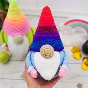 May include: A handmade crochet gnome with a rainbow-coloured hat and a beige nose. The gnome has a white beard, blue body, and pink mittens. A second gnome with a green body and rainbow hat is in the background. A rainbow and pot of gold are also visible.