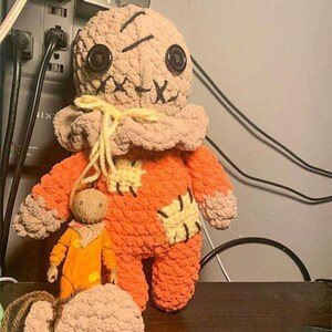 Peut inclure: Une poupée en peluche faite à la main représentant un personnage de film d'horreur. La poupée est principalement orange avec des détails beiges, des yeux en boutons noirs et des traits du visage cousus. Une figurine plus petite du même personnage est également présente.