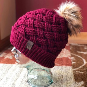 May include: A burgundy knit beanie with a woven pattern and a faux fur pom-pom. The hat features a small, brown leather tag with the initials "CBJ". The hat is displayed on a glass head.